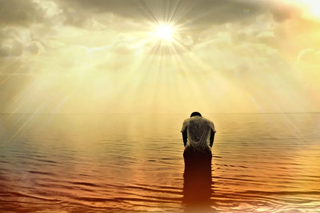 person, standing in water, head bowed, sun peaking through the clouds